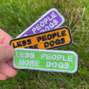 May include: Three embroidered patches with the text "LESS PEOPLE MORE DOGS". The patches are rectangular with rounded corners and feature the text in black on a purple, orange, and green background. The patches are held against a background of green grass.