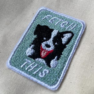 May include: Embroidered patch with a black and white dog design and a red tongue, on a light green background. The patch reads "FETCH THIS" in white cursive lettering, with a light blue border.