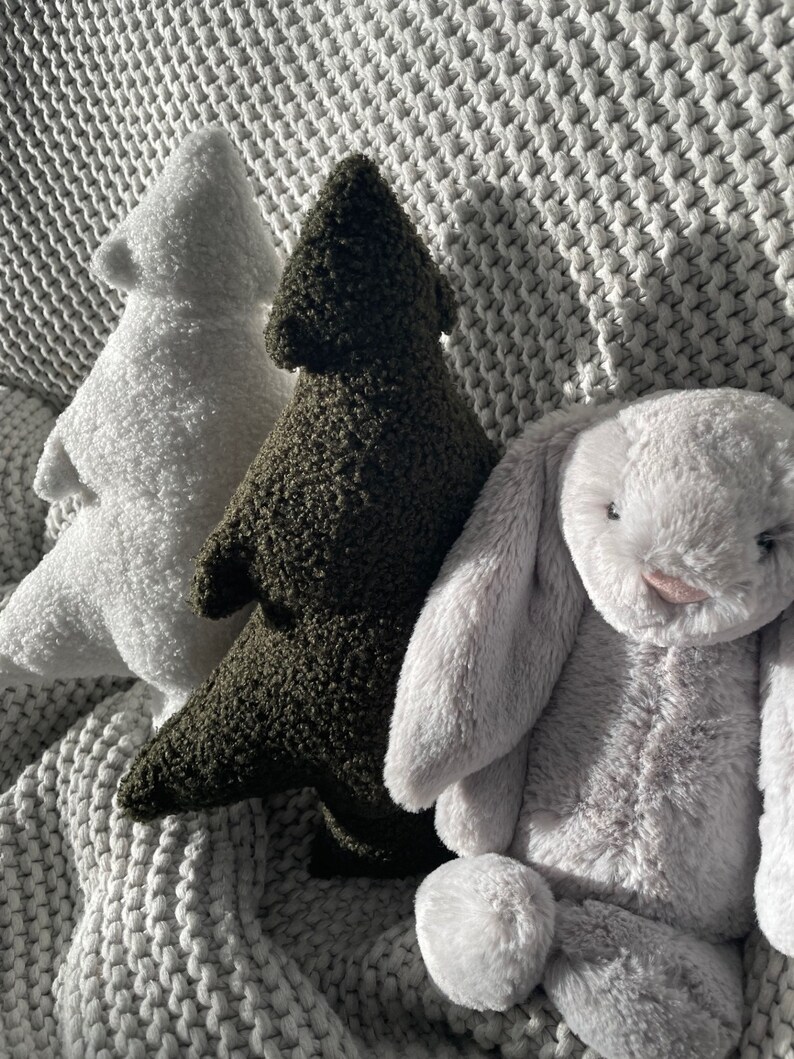 May include: Three plush toys: a white tree, a dark green tree, and a gray bunny. The toys are made of soft, textured material and are arranged on a gray, knitted blanket. The bunny has long, floppy ears and a pink nose.