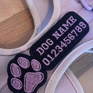 Op de afbeelding: Een lila hondentuigje met een zwart label geborduurd met "DOG NAME" en een telefoonnummer in lila. Een bijpassende pootafdrukpatch is ook bevestigd. Het tuigje is gemaakt van een zacht, duurzaam materiaal.