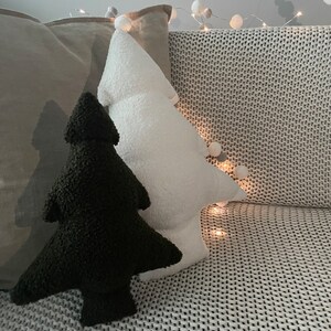 May include: Two Christmas tree-shaped pillows, one black and one white, rest on a gray textured surface. The white pillow has a pom-pom at the top. A string of lights with small white balls is in the background.