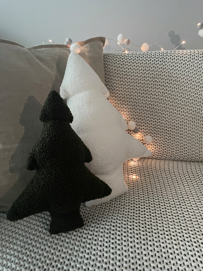 May include: Two Christmas tree-shaped pillows, one black and one white, rest on a gray textured surface. The white pillow has a pom-pom at the top. A string of lights with small white balls is in the background.