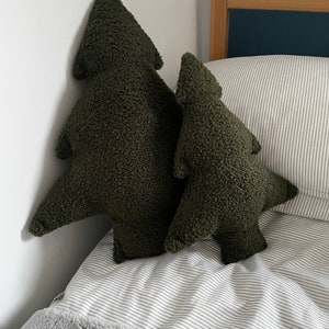 May include: Two olive green tree-shaped decorative pillows. The pillows have a textured, plush appearance and are placed on a bed with a striped comforter and a gray blanket. The pillows are different sizes.