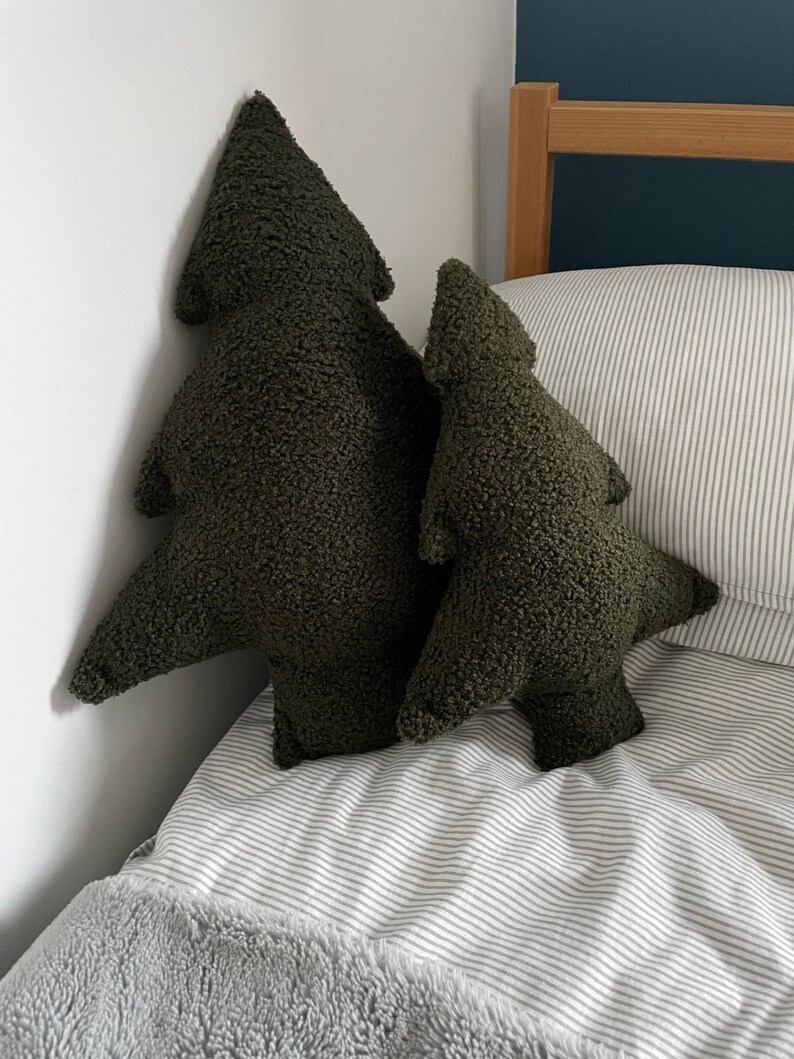 May include: Two olive green tree-shaped decorative pillows. The pillows have a textured, plush appearance and are placed on a bed with a striped comforter and a gray blanket. The pillows are different sizes.