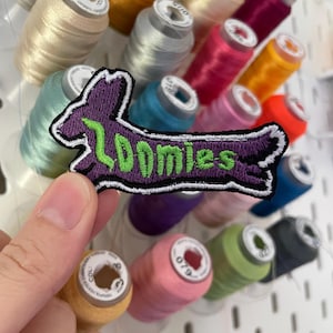 May include: A purple embroidered patch in the shape of a running rabbit with the word "Zoomies" in green. The patch has a white border. Spools of colourful thread are in the background.