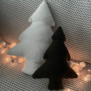 May include: Two plush Christmas tree-shaped pillows, one white and one dark green, rest on a gray knitted blanket. The pillows have a soft, textured appearance. A string of fairy lights with small, round bulbs adds a festive touch.