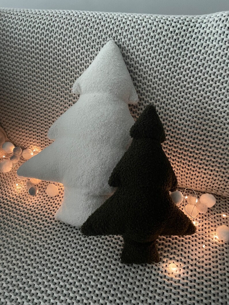 May include: Two plush Christmas tree-shaped pillows, one white and one dark green, rest on a gray knitted blanket. The pillows have a soft, textured appearance. A string of fairy lights with small, round bulbs adds a festive touch.