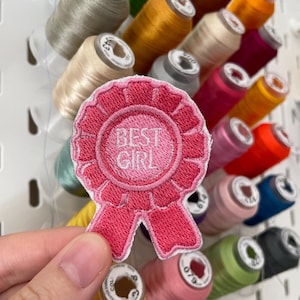 Könnte beinhalten: Ein rosa bestickter Aufnäher mit den weißen Worten "BEST GIRL". Der Aufnäher hat die Form eines Bandes und wird vor einem Hintergrund aus bunten Garnrollen gehalten.