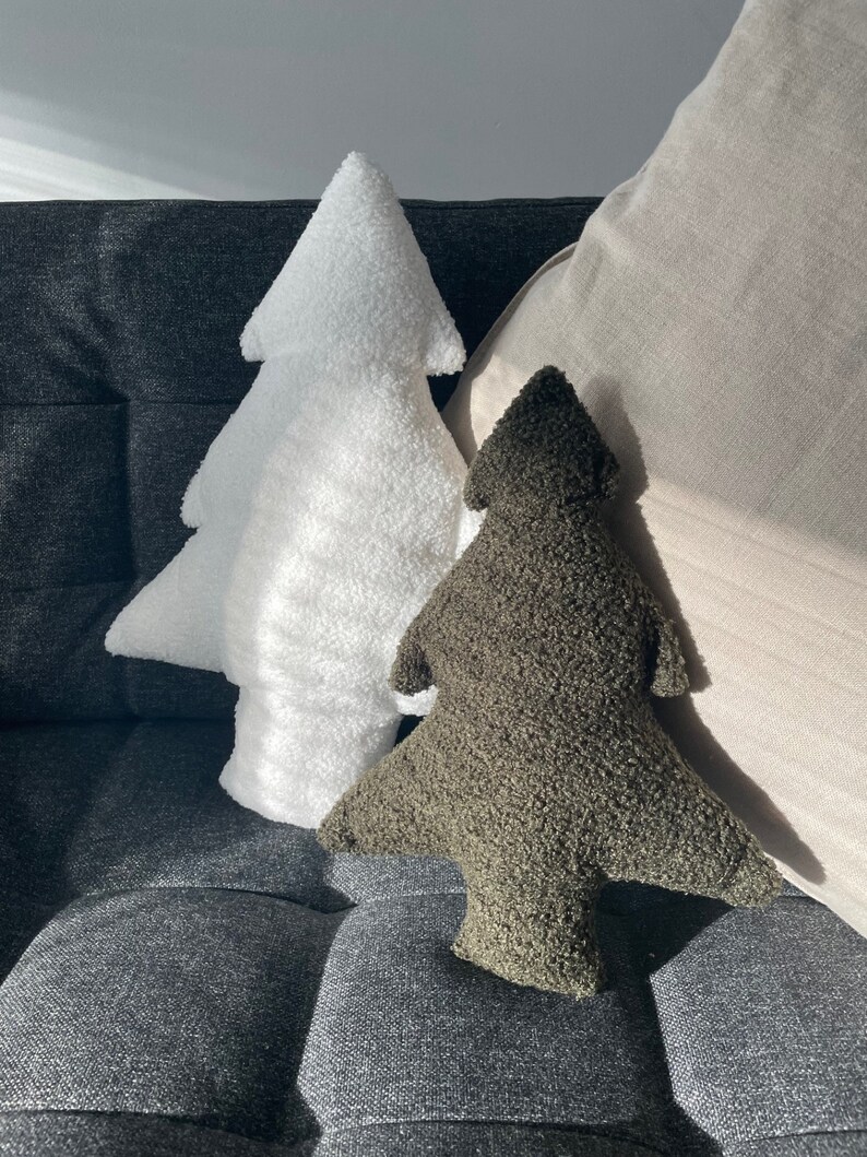 May include: Two tree-shaped decorative pillows. One is white, the other is olive green. Both pillows have a textured, plush appearance. They are resting on a dark gray upholstered surface, with a light-colored pillow in the background.