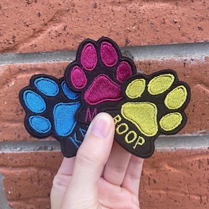 May include: Three embroidered paw print patches in blue, magenta, and yellow. Each patch has a black background and the words "KA" and "ROOP" are embroidered on two of the patches. The patches are held in a hand.