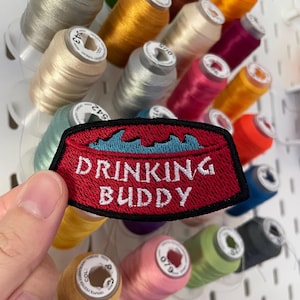 Op de afbeelding: Een rood geborduurd embleem met de witte woorden "DRINKING BUDDY" boven een blauw golfpatroon. Het embleem wordt getoond voor een display van kleurrijke klosjes garen, waaronder tinten geel, roze en groen.