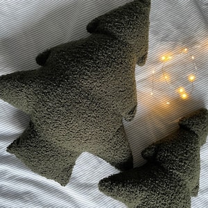 May include: Two olive green tree-shaped decorative pillows with a textured, plush surface. The pillows are different sizes and are placed on a striped, light-colored fabric. A string of small, warm white lights is in the background.