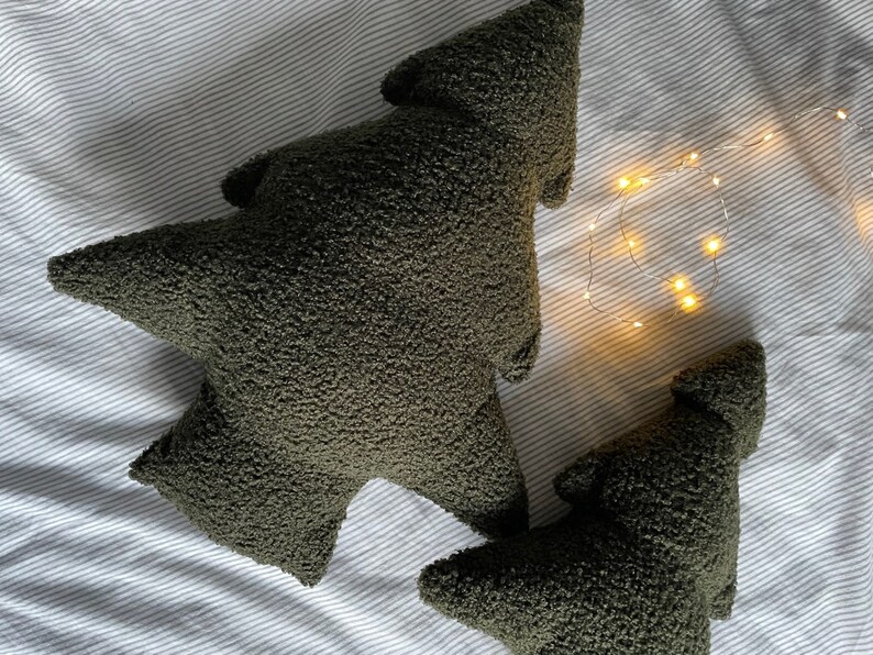 May include: Two olive green tree-shaped decorative pillows with a textured, plush surface. The pillows are different sizes and are placed on a striped, light-colored fabric. A string of small, warm white lights is in the background.
