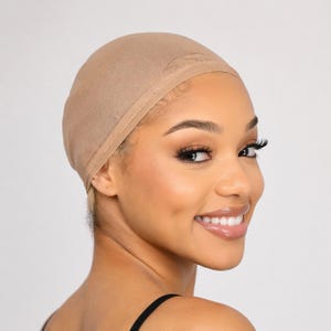 Puede incluir: Una gorra de peluca beige hecha de un material de malla fina. La gorra está diseñada para usarse debajo de una peluca para asegurar el cabello natural y proteger el cuero cabelludo. La gorra se muestra en una persona sonriente.