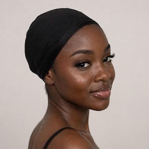 Puede incluir: Una cubierta para la cabeza negra, probablemente un gorro para dormir o un protector para el cabello, es usado por una persona de piel oscura. La gorra está hecha de una tela suave y mate. La persona mira hacia la cámara.