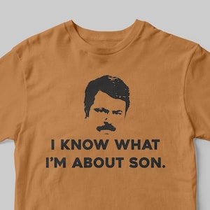May include: A brown t-shirt featuring a black silhouette of Ron Swanson from the show Parks and Recreation. The text "I know what I'm about son." is printed on the shirt.