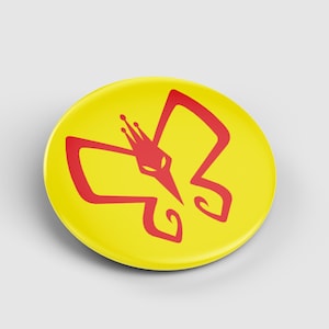 May include: A yellow button with a red butterfly design. The butterfly has a crown on its head. The button is round and has a glossy finish. The design is simple and bold.