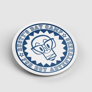 Könnte beinhalten: Ein weiß-blauer runder Button mit dem Text "Rusty's Day Camp For Boy Adventurers". Der Button zeigt eine Cartoon-Glühbirne mit Brille. Das Design ist von einem blauen Sternenmuster umgeben.