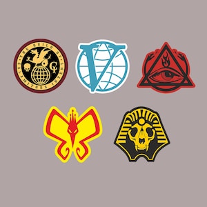 Puede incluir: Cinco pegatinas coloridas con diferentes diseños. La primera pegatina es un círculo rojo con un globo dorado y un pájaro con una corona. La segunda pegatina es un círculo azul con una letra V blanca y un globo blanco. La tercera pegatina es un triángulo rojo con un ojo negro y una llama. La cuarta pegatina es una mariposa amarilla con una corona roja y una cara roja. La quinta pegatina es una cabeza de faraón negra con una corona amarilla y una calavera amarilla.