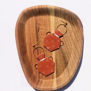 Könnte beinhalten: Ein Paar ockerfarbene Ohrringe mit geometrischem Design und goldfarbenen Reifen. Die Ohrringe sind wie kleine Vasen mit weißen Punktmustern geformt. Sie werden auf einem ovalen Holztablett präsentiert.