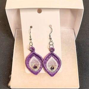 May include: A pair of embroidered purple and white teardrop earrings with silver-tone hooks. The earrings are displayed in a white gift box. The earrings have a detailed floral design with a small, dark bead in the center.