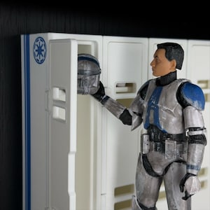 May include: A detailed figurine of a soldier in a futuristic uniform, holding a helmet near a white locker. The uniform is primarily silver with blue accents on the shoulders and chest. The locker has a blue and white design.