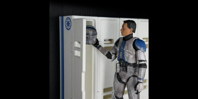 May include: A detailed figurine of a soldier in a futuristic uniform, holding a helmet near a white locker. The uniform is primarily silver with blue accents on the shoulders and chest. The locker has a blue and white design.