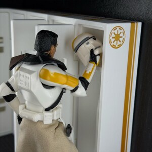 May include: A Star Wars action figure in a white and yellow uniform is placing a helmet into a white locker. The locker has gold stripes and a gold emblem. The figure is wearing a tan skirt.
