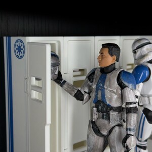 May include: Action figures of Star Wars clone troopers. One figure is holding a helmet in front of a white locker with blue accents. Another trooper stands nearby. The figures are in silver, white, and blue armor.
