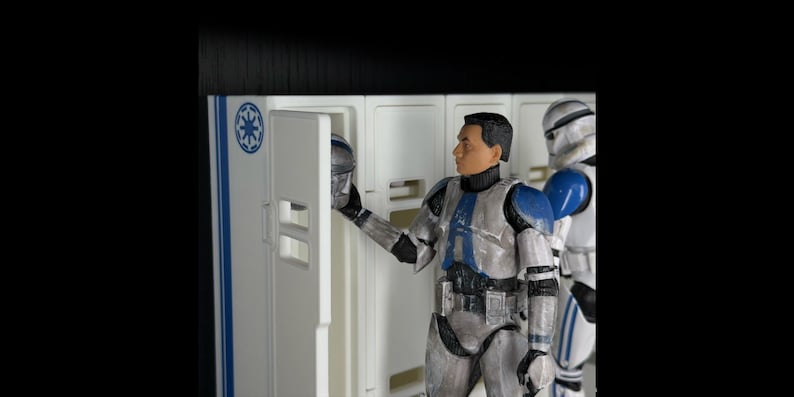 May include: Action figures of Star Wars clone troopers. One figure is holding a helmet in front of a white locker with blue accents. Another trooper stands nearby. The figures are in silver, white, and blue armor.