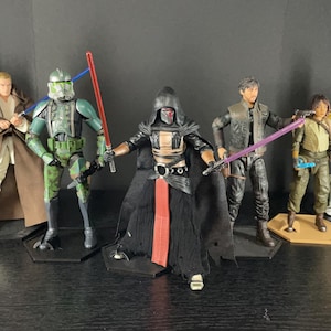 May include: A collection of Star Wars action figures, including a green and black clone trooper, a black-robed figure with a red sash, a brown-robed figure, a blue and white clone trooper, a gold C-3PO, a black-clad figure with a purple lightsaber, a brown-clad figure with a blue lightsaber, and a brown-clad figure with a blue lightsaber.