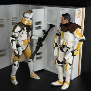 May include: Two action figures of Star Wars clone troopers in white, yellow, and brown armor stand near a white locker. One figure holds a black blaster rifle. The figures have helmets and are posed in a scene.