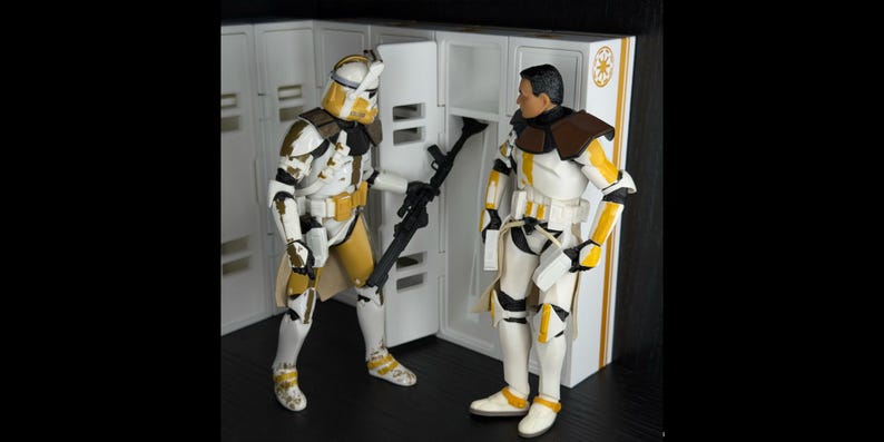 May include: Two action figures of Star Wars clone troopers in white, yellow, and brown armor stand near a white locker. One figure holds a black blaster rifle. The figures have helmets and are posed in a scene.
