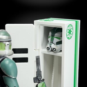 May include: A white and green display case with a Star Wars helmet inside. The case has green stripes and a green emblem. A figure in green and white armor stands to the left, holding a blaster.