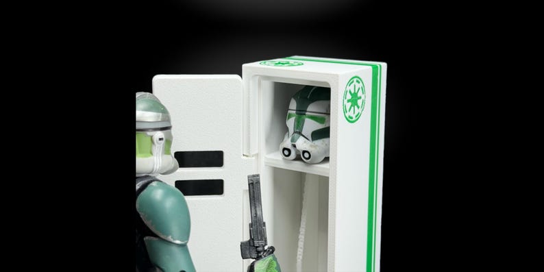 May include: A white and green display case with a Star Wars helmet inside. The case has green stripes and a green emblem. A figure in green and white armor stands to the left, holding a blaster.