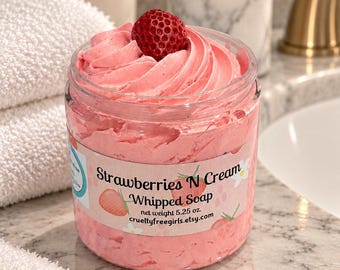 Op de afbeelding: Een doorzichtige plastic pot gevuld met roze opgeklopte zeep, bekroond met een enkele rode aardbei. Het etiket vermeldt Strawberries N Cream Whipped Soap, net weight 5.25 oz. en crueltyfreegirls.etsy.com, met illustraties van aardbeien en witte bloemen.