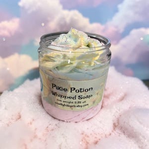 May include: A clear plastic jar filled with pastel-colored whipped soap topped with glitter. The label reads Pixie Potion Whipped Soap, net weight 5.25 oz., and crueltyfreegirls.etsy.com. The soap features swirls of light blue, yellow, and pink.