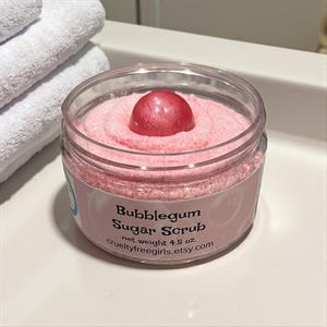 May include: A clear jar filled with pink bubblegum sugar scrub, topped with a shiny, round, pink embellishment. The jar has a label with the text "Bubblegum Sugar Scrub" and "crueltyfreegirls.etsy.com". The net weight is 4.5 oz.