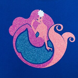 Op de afbeelding: Een roze en blauwe glitter zeemeermin ontwerp op een koninklijk blauwe t-shirt. De zeemeermin heeft een bloem in haar haar en een glinsterende staart.