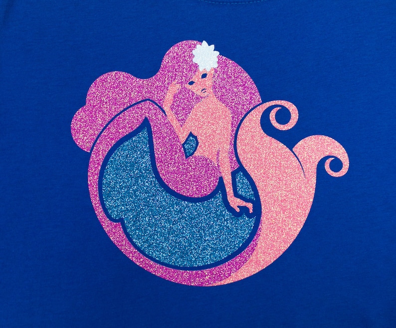 Op de afbeelding: Een roze en blauwe glitter zeemeermin ontwerp op een koninklijk blauwe t-shirt. De zeemeermin heeft een bloem in haar haar en een glinsterende staart.