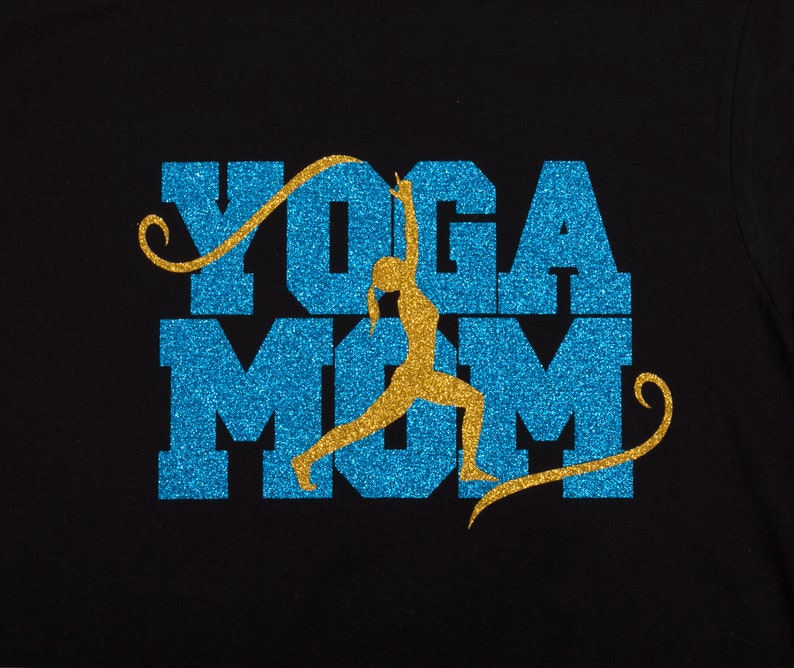 Op de afbeelding: Een zwart T-shirt met een blauw en gouden glitterontwerp met de tekst "Yoga Mom" en een silhouet van een vrouw die yoga doet in het midden van het woord "Mom".