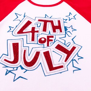 May include: A white raglan shirt with red sleeves featuring a glittery red and blue graphic that reads 4TH OF JULY. The text is surrounded by blue glitter star outlines and has a silver glitter border, creating a festive and bold appearance.