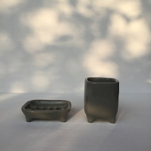 Puede incluir: Un juego de dos accesorios de baño de cerámica gris. El jabonera tiene cuatro patas y una forma rectangular. El portacepillos de dientes es más alto y tiene una forma cuadrada con cuatro patas.