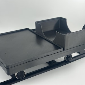 May include: A black 3D-printed trolley featuring a flat rectangular platform and a raised storage compartment with a curved cutout. The trolley is mounted on four small wheels and is positioned on a black track system, designed for smooth movement.