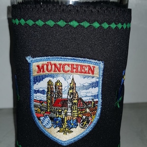 Peut inclure: Un manchon de bière en néoprène noir avec des coutures vertes et un écusson brodé blanc, bleu et rouge qui dit "MÜNCHEN" avec une représentation d'un horizon urbain.