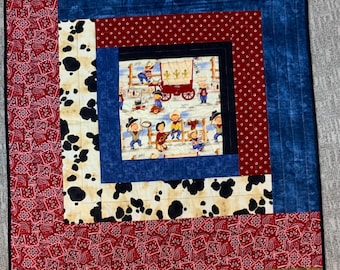May include: A quilt with a patchwork design featuring cowboys, wagons, and paisley patterns. The quilt is made with a variety of fabrics in red, blue, white, and black.