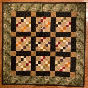 May include: Square quilt with a patchwork design. The quilt features nine squares with a central pattern of colorful squares surrounded by black borders. The edges have a floral pattern in green and brown.