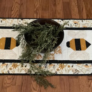 May include: A white table runner with a black border and a floral pattern featuring bees. The runner has two black and yellow bee appliques in the center. A brown bowl with green foliage is on top of the runner.