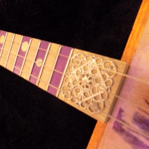 Op de afbeelding: Een gitaarhals met een toets van licht hout, voorzien van paarse rechthoekige inlays en ronde gele markeringen. Een zilverkleurig, ingewikkeld, vierkant metalen filigraan ornament is bevestigd aan de basis van de toets.