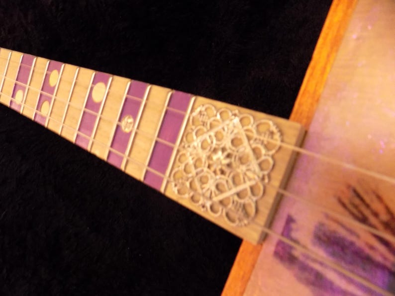 Op de afbeelding: Een gitaarhals met een toets van licht hout, voorzien van paarse rechthoekige inlays en ronde gele markeringen. Een zilverkleurig, ingewikkeld, vierkant metalen filigraan ornament is bevestigd aan de basis van de toets.